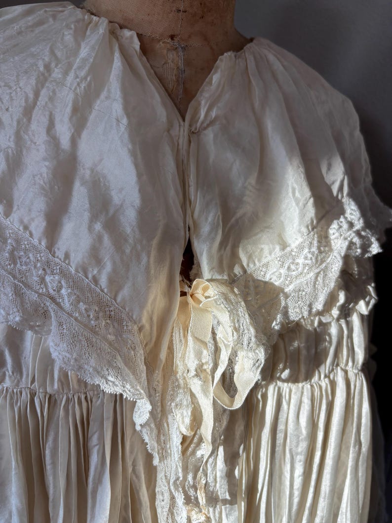 May include: Cream-colored vintage dress with a V-neckline and delicate lace trim. The dress features a flowing, layered design with a ribbon detail at the waist. The fabric appears to be silk or a similar lightweight material.