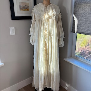 May include: A vintage cream-colored dressing gown with a ruffled collar and sleeves, displayed on a mannequin. The long, flowing gown features lace trim and a delicate, textured fabric. The gown is floor-length and has a button-up front.