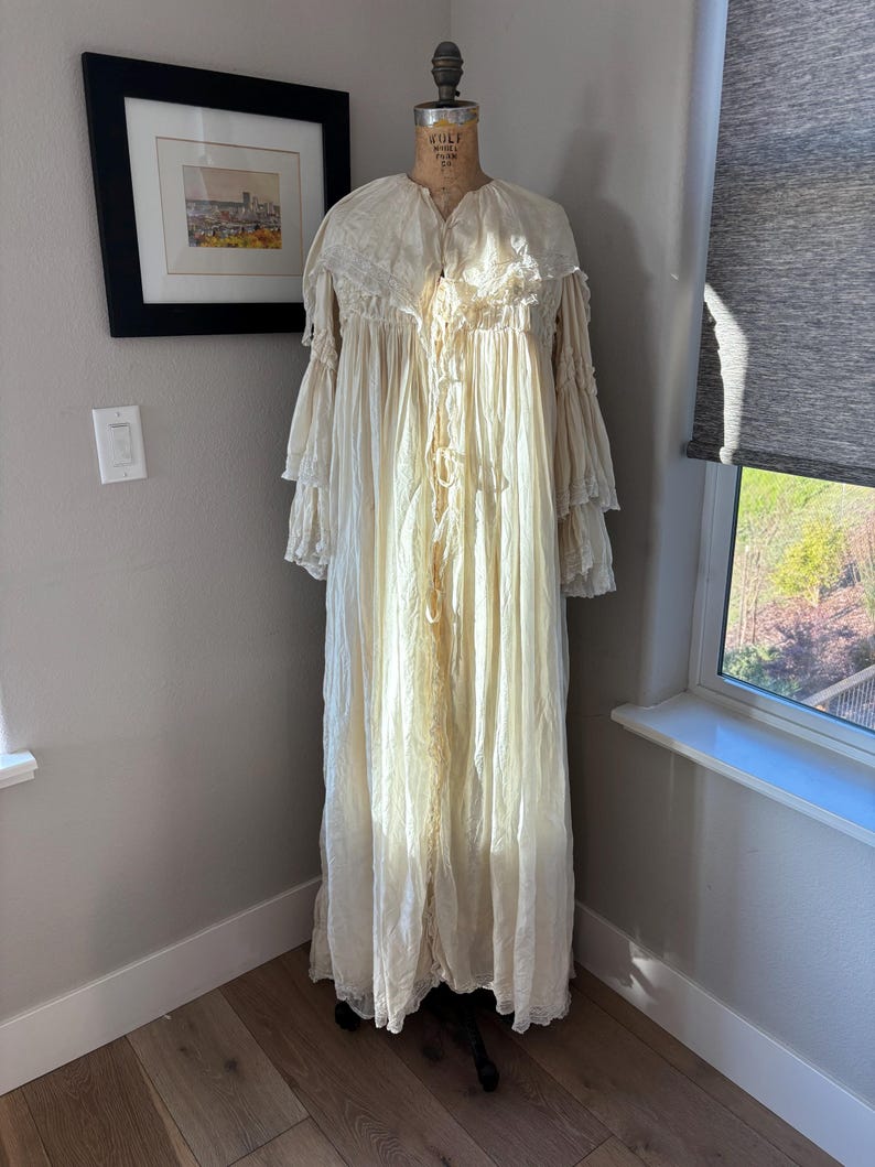 May include: A vintage cream-colored dressing gown with a ruffled collar and sleeves, displayed on a mannequin. The long, flowing gown features lace trim and a delicate, textured fabric. The gown is floor-length and has a button-up front.