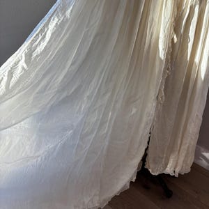 May include: A flowing, off-white vintage gown with a delicate lace trim along the hemline. The fabric appears lightweight and semi-sheer, with soft folds and draping. The gown is displayed on a dark stand, suggesting a focus on the garment's texture and design.