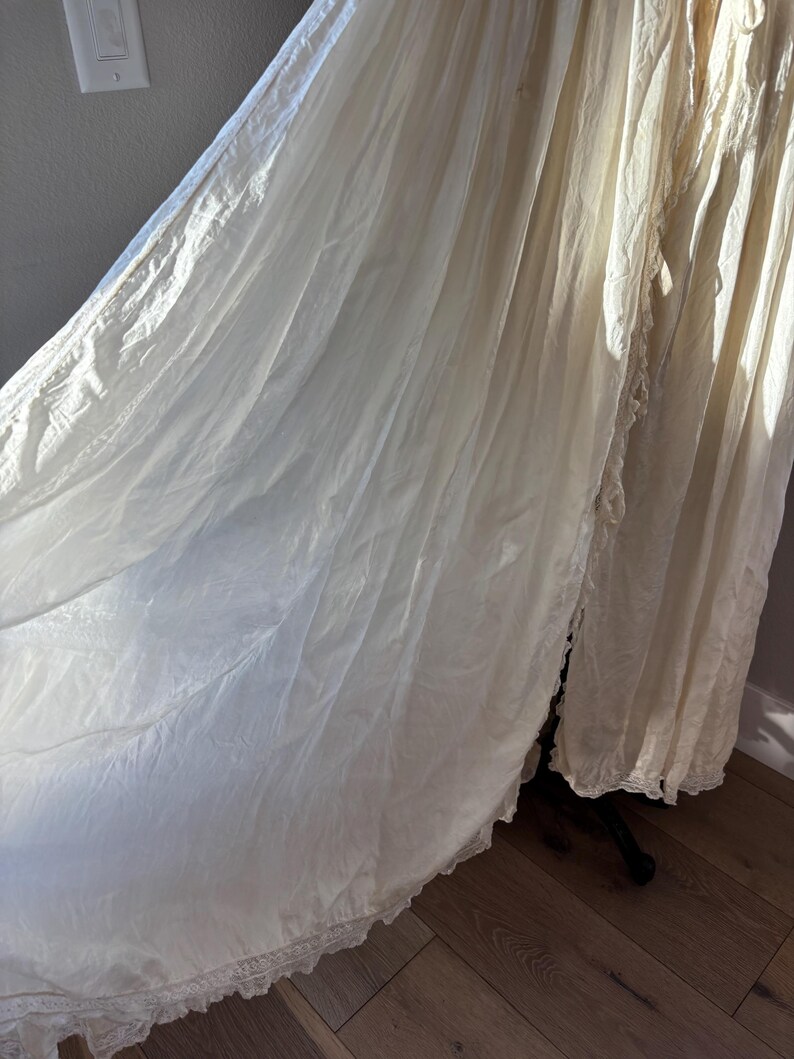 May include: A flowing, off-white vintage gown with a delicate lace trim along the hemline. The fabric appears lightweight and semi-sheer, with soft folds and draping. The gown is displayed on a dark stand, suggesting a focus on the garment's texture and design.