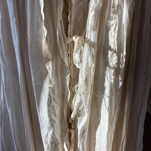 May include: Close-up of off-white fabric with a layered, textured appearance. The fabric has a flowing, draped quality with visible seams and delicate, frayed edges. The color is a soft, creamy white, with shadows adding depth and dimension to the folds.