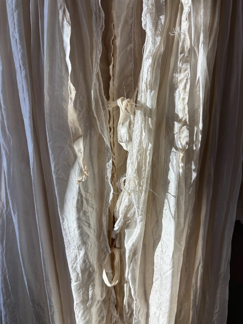 May include: Close-up of off-white fabric with a layered, textured appearance. The fabric has a flowing, draped quality with visible seams and delicate, frayed edges. The color is a soft, creamy white, with shadows adding depth and dimension to the folds.