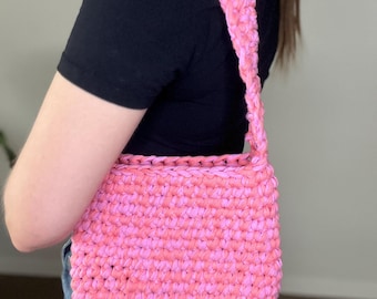 May include: A pink crocheted shoulder bag with a long strap. The bag is made of a soft, textured yarn.