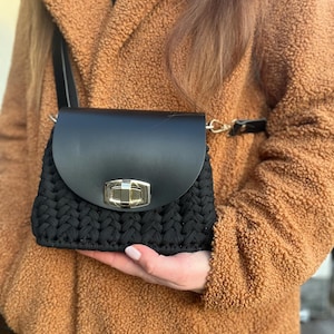 May include: A black crocheted crossbody bag with a black leather flap and gold clasp. The bag is being held by a person wearing a brown fuzzy coat.