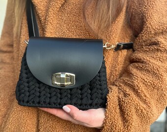 May include: A black crocheted crossbody bag with a black leather flap and gold clasp. The bag is being held by a person wearing a brown fuzzy coat.