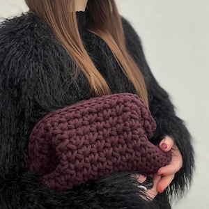 May include: A burgundy crocheted makeup bag with a textured pattern. The bag is held in the hands of a person wearing a black fuzzy jacket.