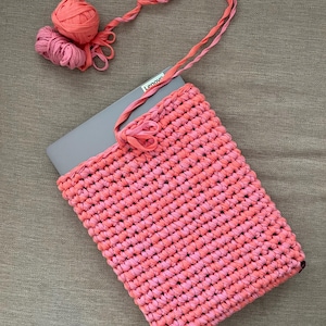 May include: A pink and coral crocheted laptop sleeve with a long pink and coral crocheted strap. The sleeve is lying on a beige surface with a laptop inside. The strap is looped over the laptop and the ends of the strap are hanging down. There is a ball of pink and coral yarn next to the sleeve.