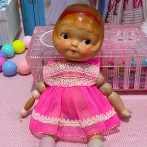 Op de afbeelding: Een vintage pop met een geschilderd gezicht, blauwe ogen en een rode haarband. De pop draagt een felroze jurk met witte kanten rand en rode schoenen. De armen en benen van de pop zijn beweeglijk.