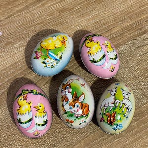 May include: Five vintage Easter egg-shaped tins with different pastel colored backgrounds and illustrations. The tins feature chicks, bunnies, and lambs.