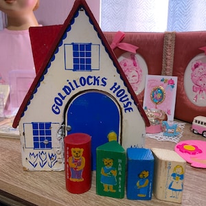Puede incluir: Una casa de madera para juguetes con una puerta y ventanas azules, pintada con las palabras "Goldilocks House". Hay cuatro bloques de madera con osos en ellos, etiquetados como "Papa Bear", "Mama Bear", "Baby Bear" y "Goldilocks".