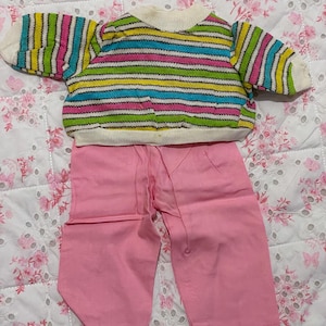 Könnte beinhalten: Ein Puppenoutfit mit einem gestreiften Pullover und einer rosa Hose. Der Pullover hat horizontale Streifen in verschiedenen Farben, darunter Rosa, Blau, Gelb und Grün. Die Hose ist einfarbig hellrosa. Das Outfit ist für eine kleine Puppe.