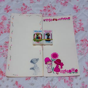 May include: Two rectangular off-white stationery sheets and two small rectangular erasers. The left sheet features a small sketch of a child, while the right sheet displays a colorful illustration of two children and flowers. The erasers are labeled PIPO & PAPO.