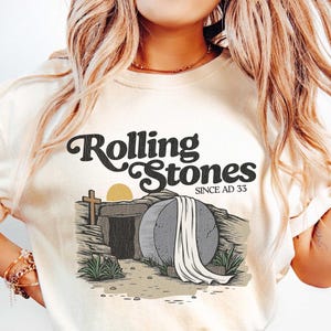 May include: A white t-shirt with a graphic design of a stone tomb with a white sheet draped over it. The text "Rolling Stones Since AD 33" is above the tomb.