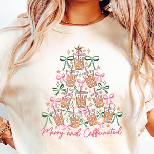 May include: Cream-colored t-shirt featuring a Christmas tree design made of iced coffee cups, bows, and stars. The phrase "Merry and Caffeinated" is written in pink script at the bottom. A festive and fun design.