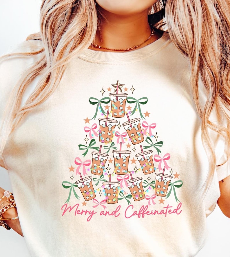 May include: Cream-colored t-shirt featuring a Christmas tree design made of iced coffee cups, bows, and stars. The phrase "Merry and Caffeinated" is written in pink script at the bottom. A festive and fun design.