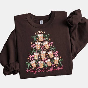 May include: A dark brown sweatshirt with a Christmas tree design made of coffee cups, bows, and lights. The text "Merry and Caffeinated" is written in pink script at the bottom of the tree.