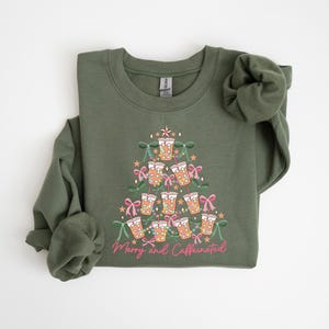 May include: Olive green sweatshirt with a graphic of a Christmas tree made of coffee cups, bows, and stars. The text "Merry and Caffeinated" is printed in pink script at the bottom of the design. The sweatshirt has long sleeves.