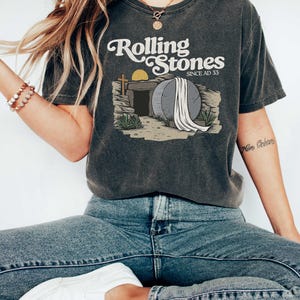 May include: A dark grey t-shirt with a vintage-style graphic of a tombstone with a white sheet draped over it. The text "Rolling Stones Since AD 33" is printed above the graphic.