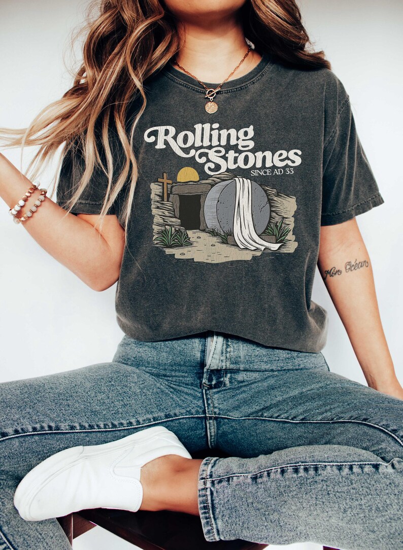 May include: A dark grey t-shirt with a vintage-style graphic of a tombstone with a white sheet draped over it. The text "Rolling Stones Since AD 33" is printed above the graphic.