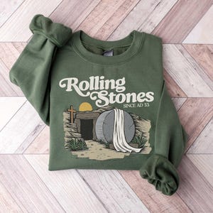 May include: Green sweatshirt with a graphic of a stone tomb with a white cloth draped over it. The text "Rolling Stones Since AD 33" is printed on the sweatshirt.