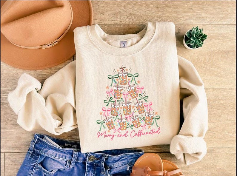 May include: Cream-colored sweatshirt with a Christmas tree design made of iced coffee cups, bows, and stars. The text "Merry and Caffeinated" is written in pink script. The sweatshirt is paired with blue jeans, sandals, and a brown hat.