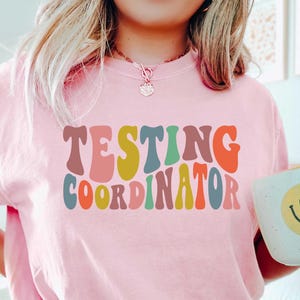 May include: Pink t-shirt with the text "TESTING COORDINATOR" in a retro, colorful font.