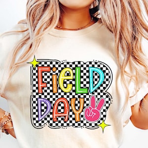 May include: A white t-shirt with a colorful graphic that says "Field Day" in a checkered pattern with a pink peace sign.
