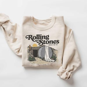May include: A beige sweatshirt with a graphic print of a stone tomb with a white sheet on top. The text "Rolling Stones Since AD 33" is printed above the image.