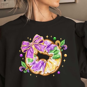May include: Black sweatshirt with a purple and green donut design. The donut is decorated with purple and gold sprinkles and a purple bow. The donut is surrounded by green and gold leaves and purple beads.