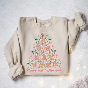 May include: Beige sweatshirt with a Christmas tree design made of iced coffee cups, bows, and stars. The phrase "Merry and Caffeinated" is written in pink script at the bottom. A festive and cozy holiday sweater.