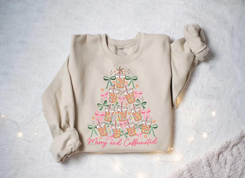 May include: Beige sweatshirt with a Christmas tree design made of iced coffee cups, bows, and stars. The phrase "Merry and Caffeinated" is written in pink script at the bottom. A festive and cozy holiday sweater.