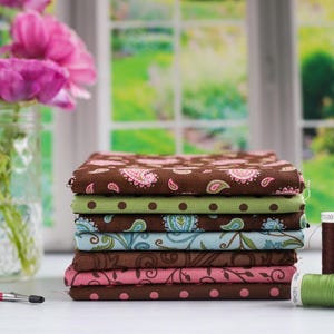 May include: A stack of eight fabric squares in various patterns, including paisley, polka dots, and floral designs. The fabrics are in shades of brown, green, pink, and blue. Three spools of thread are in the foreground, in brown, pink, and green.