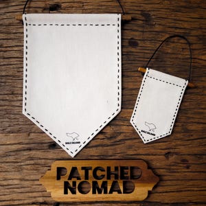 May include: Two white canvas pennant flags with black stitching and wooden dowels, one large and one small, hang against a dark wood background. The small flag has a black cord for hanging. A wooden sign below reads "PATCHED NOMAD."