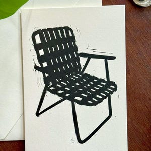 Może przedstawiać: Czarno-biały linoryt przedstawiający tkany leżak ogrodowy na białej karcie. Krzesło ma prostokątne oparcie i siedzisko z podłokietnikami. Karta znajduje się w białej kopercie.