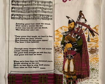May include: A white tea towel with a red, white, and blue plaid pattern. The tea towel features a picture of a man playing bagpipes in a kilt. The text "Scotland" is printed in red and white letters. The text "Amazing Grace" is printed in black letters with musical notes. The lyrics to the hymn "Amazing Grace" are printed below the musical notes.