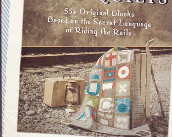 May include: The book "HOBO QUILTS" is displayed with a colorful quilt draped over a wooden crate. The quilt features various geometric shapes and patterns. A vintage suitcase and lantern are in the foreground, set against a backdrop of railroad tracks.