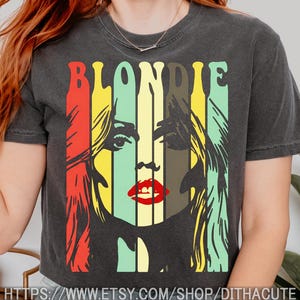 May include: A charcoal gray short-sleeve t-shirt featuring a graphic print of the word BLONDIE in a bold, retro font. The design includes vertical color stripes in red, yellow, and light green, overlaying a stylized portrait of a face with red lips.