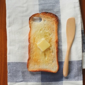 Puede incluir: Funda de teléfono con forma de rebanada de pan tostado con una nuez de mantequilla. La funda es de color marrón claro con un cuadrado de mantequilla amarilla en el centro. Una espátula de madera descansa sobre un paño a cuadros.