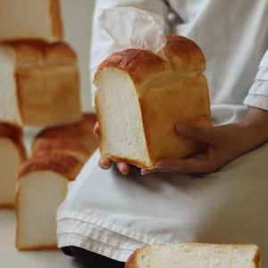 May include: A person holding a white loaf of bread in their hands. There are other loaves of bread in the background.