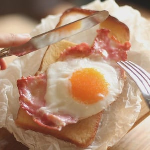 Könnte beinhalten: Ein Frühstücks-Sandwich mit Spiegelei, Speck und Toast. Das Ei hat ein leuchtend orangefarbenes Eigelb und weiße Eier. Der Speck ist rot und knusprig. Ein Messer und eine Gabel werden verwendet, um das Sandwich zu schneiden.