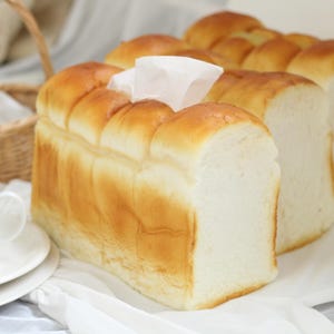 May include: A loaf of golden-brown bread with a soft, white interior. The bread is sliced and arranged on a white surface, with a white teacup and saucer in the foreground. A wicker basket is in the background.