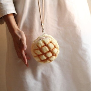 Op de afbeelding: Een ronde, lichtbruine, gebakvormige tas met een crèmekleurige band. De tas is gemaakt om eruit te zien als een broodje met een rasterpatroon.