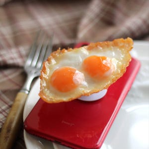 May include: A red smartphone with a white pop-up grip is topped with a fried egg with two orange yolks. A silver fork with a wooden handle rests on a white plate, with a plaid background.
