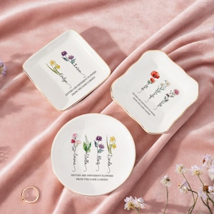 Puede incluir: Tres platos de cerámica blanca para baratijas con bordes dorados, cada uno con diseños florales y nombres. El texto "Sisters are different flowers from the same garden" está impreso en cada plato. Uno es cuadrado, otro festoneado y otro redondo.