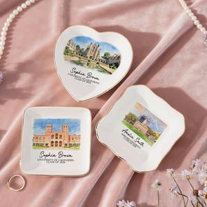 May include: Three white ceramic trinket dishes with gold rims, in heart, square, and scalloped shapes. Each dish features a watercolor image of a university building and the name and graduation year of a student. The dishes are on a pink velvet surface.