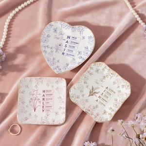 Puede incluir: Tres platos decorativos de cerámica en forma de corazón, cuadrado e irregular, con estampados florales y ribetes dorados. El plato en forma de corazón tiene un diseño floral azul, mientras que los otros tienen estampados florales dorados y rosas. Cada plato tiene texto.