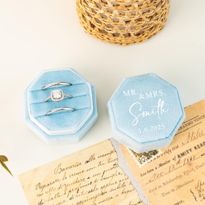 May include: Light blue velvet ring boxes with silver rings inside. One box is open, displaying three rings. The other box is closed, with "MR & MRS Smith 1.6.2025" printed on top. A woven basket is in the background.