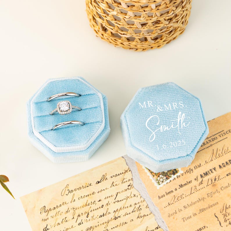 May include: Light blue velvet ring boxes with silver rings inside. One box is open, displaying three rings. The other box is closed, with "MR & MRS Smith 1.6.2025" printed on top. A woven basket is in the background.