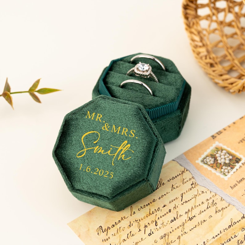 May include: A dark green velvet ring box with gold lettering reading "MR. & MRS. Smith 1.6.2025". The box is open, displaying three rings. A woven basket and a handwritten note are visible in the background.